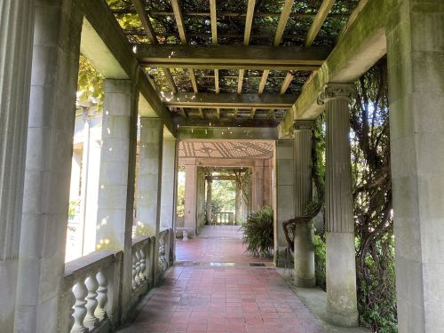 A stone walkway with columns and a trellis, surrounded by greenery and soft sunlight filtering through the leaves.