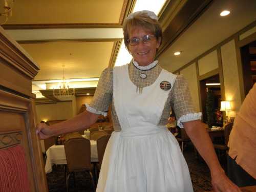 A smiling woman in a vintage-style waitress uniform stands in a dining area, with tables set in the background.