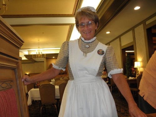 A smiling woman in a vintage-style waitress uniform stands in a dining area, with tables set in the background.