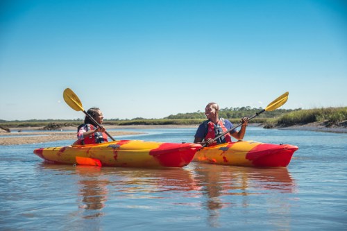 Two people in yellow kayaks paddle together on a calm waterway under a clear blue sky.
