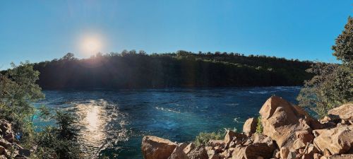 A panoramic view of a river surrounded by trees, with the sun shining brightly above the horizon.