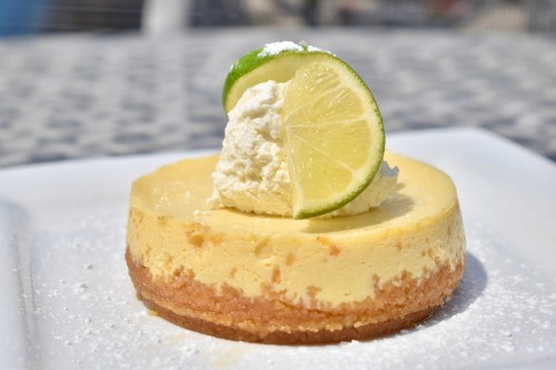 A slice of cheesecake topped with whipped cream and a lime wedge, served on a white plate.