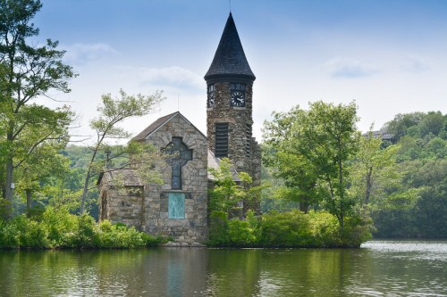 A stone church with a clock tower surrounded by lush greenery and reflecting in a calm lake under a blue sky.