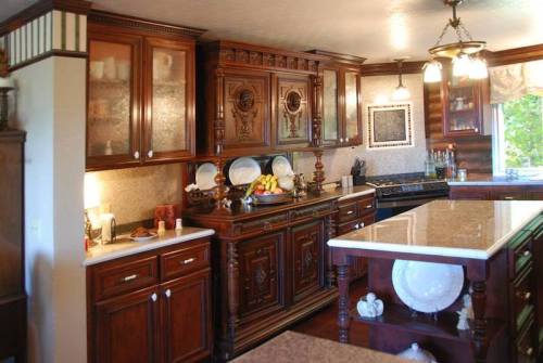A warm, wooden kitchen featuring ornate cabinets, a marble island, and decorative dishware on display.
