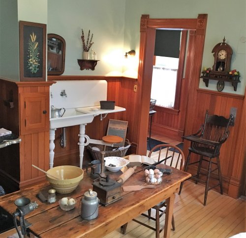 A vintage kitchen with wooden walls, a table set with cooking utensils, and a cozy dining area in the background.