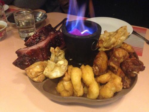 A platter of assorted fried foods, including wings and dumplings, with a small pot of flaming sauce in the center.