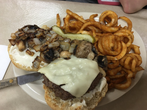 A plate with a burger topped with melted cheese, sautéed onions, and mushrooms, served with curly fries and a pickle.