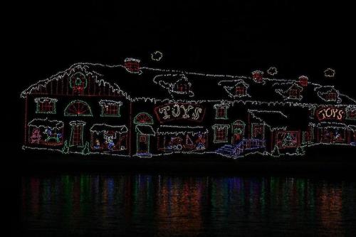 A brightly lit building decorated with colorful Christmas lights, reflecting on dark water. Snowy roof and festive details.