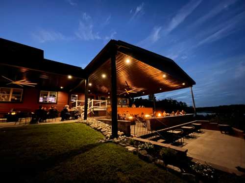A cozy restaurant patio illuminated at night, with diners enjoying the view and a serene lake in the background.