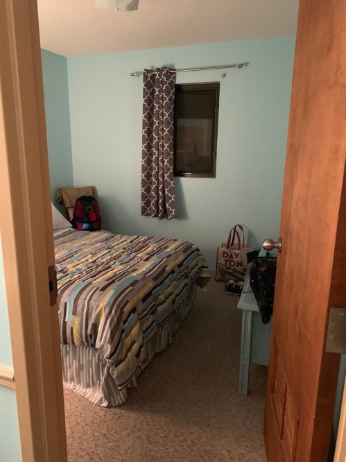 A small bedroom with a bed, light blue walls, a window with curtains, and a door partially open.