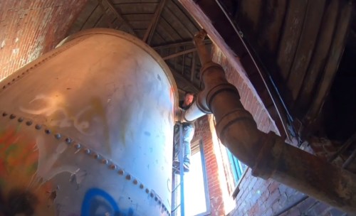 A person climbs on a large metal tank inside a brick building, with pipes and graffiti visible around them.