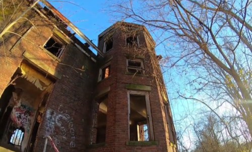 A tall, abandoned brick building with broken windows, surrounded by trees under a clear blue sky.