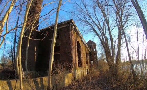 Abandoned brick building surrounded by bare trees, with a lake visible in the background under a clear blue sky.