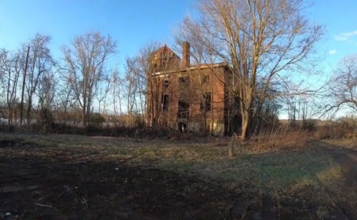An abandoned brick building surrounded by bare trees and overgrown grass under a clear blue sky.