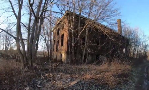 An abandoned brick building surrounded by trees and overgrown vegetation, with a clear blue sky in the background.