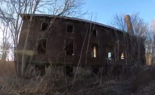 Abandoned brick building surrounded by overgrown vegetation and trees, with several broken windows.