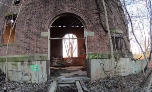 An abandoned brick building with a large arched doorway, surrounded by overgrown vegetation and graffiti.