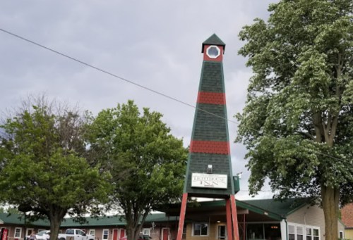 A tall, green and red clock tower stands in front of a building surrounded by trees under a cloudy sky.