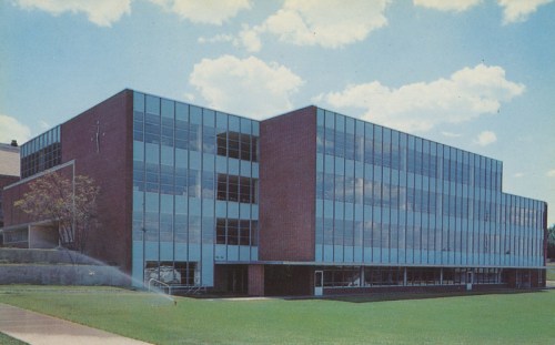 Modern brick building with large glass windows, set against a blue sky with fluffy clouds. Green lawn in the foreground.