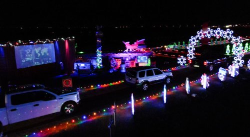 A festive display of colorful holiday lights and decorations along a driveway, with vehicles parked nearby at night.