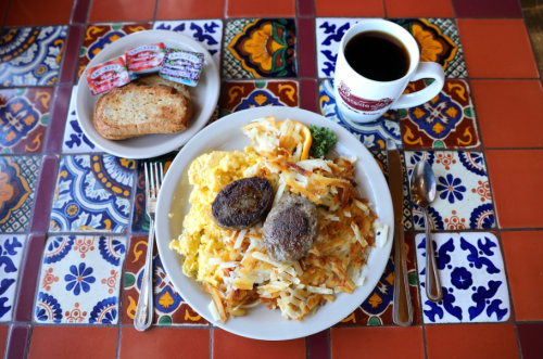 A hearty breakfast plate with scrambled eggs, hash browns, sausage patties, toast, and coffee on a colorful tiled table.