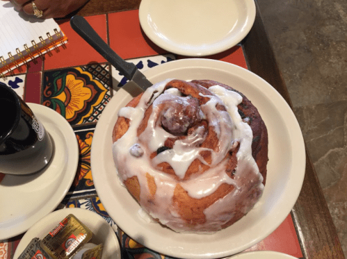 A large cinnamon roll drizzled with icing, placed on a decorative plate with a coffee cup and butter packets nearby.