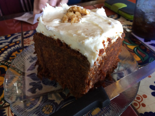 A large square slice of carrot cake topped with cream cheese frosting and a walnut, on a decorative plate.