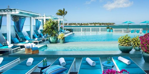 A serene poolside scene with lounge chairs, vibrant plants, and umbrellas, overlooking a calm ocean under a clear blue sky.