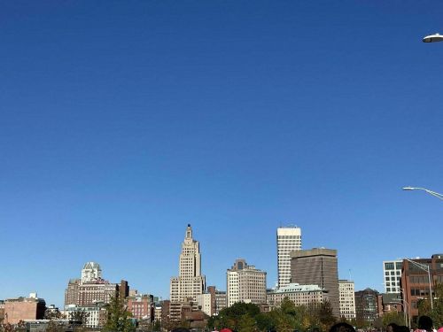 City skyline under a clear blue sky, featuring various tall buildings and a hint of greenery in the foreground.
