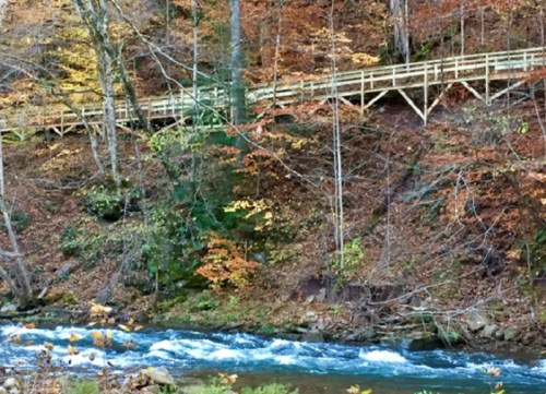 A serene river flows beside a wooden walkway, surrounded by vibrant autumn foliage and trees.