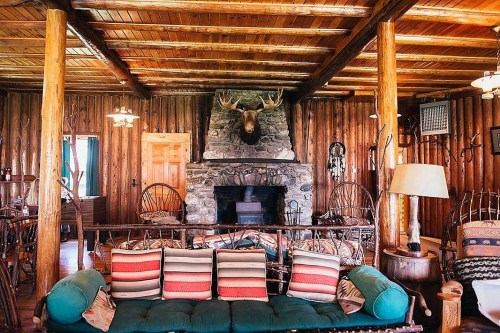 Cozy rustic living room with wooden beams, stone fireplace, and a mounted deer head, featuring a green sofa and striped cushions.