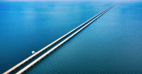Aerial view of a long, straight bridge extending over calm blue waters, creating a striking horizon line.