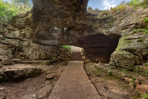 A stone pathway leads through a large rock archway, surrounded by greenery and steps ascending in the background.