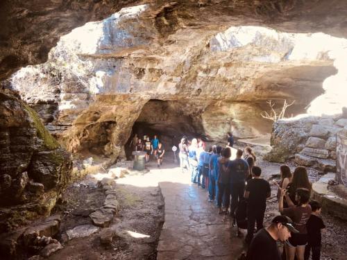 A group of people stands in line inside a large cave, with natural rock formations and light streaming in from above.