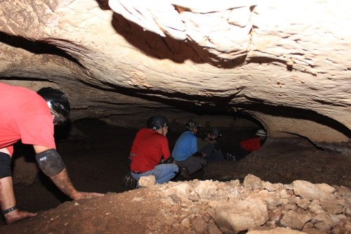 A group of cavers in helmets exploring a narrow cave passage with rocky walls and a low ceiling.
