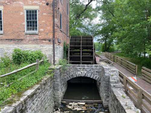 A historic waterwheel beside a stone structure, surrounded by greenery and a calm waterway.