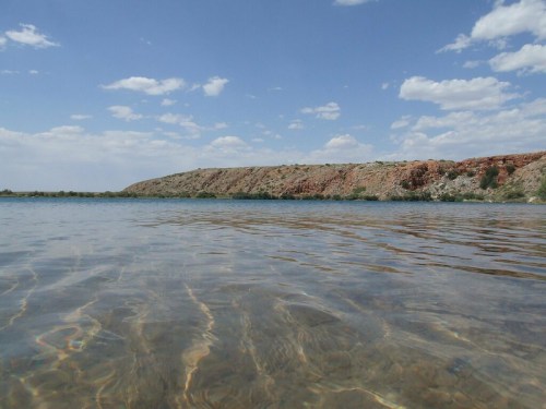 A serene view of clear water with a rocky shoreline and blue sky dotted with clouds in the background.
