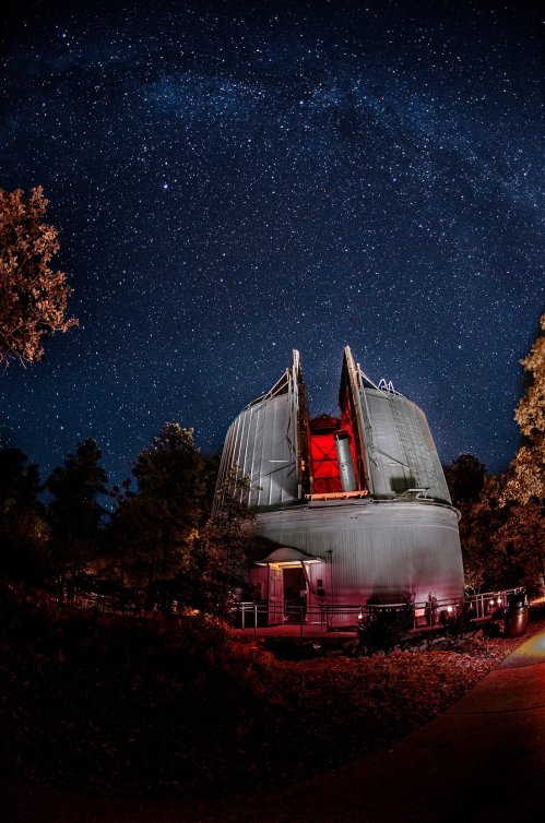 A telescope observatory under a starry night sky, illuminated by red lights, surrounded by trees.