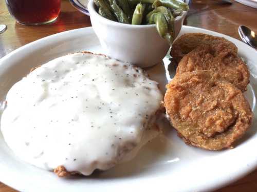 A plate featuring a breaded chicken patty smothered in white gravy, fried green tomatoes, and a side of green beans.