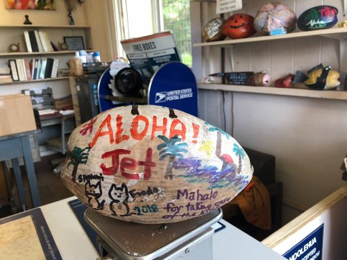 A painted rock with "Aloha! Jet" and colorful designs, displayed in a postal office with other decorated rocks in the background.