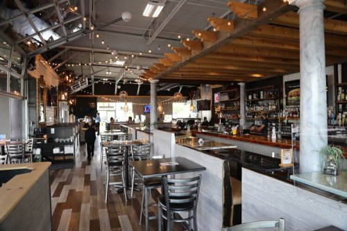 A modern restaurant interior featuring a bar, wooden beams, and string lights, with tables and chairs arranged for dining.