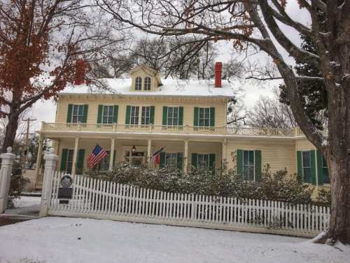 A large yellow house with green shutters, snow-covered yard, and a white picket fence, surrounded by bare trees.