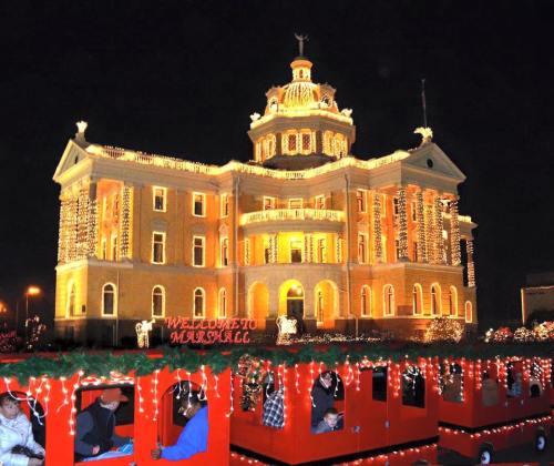 A beautifully lit historic building at night, adorned with festive lights, with a decorated train in the foreground.