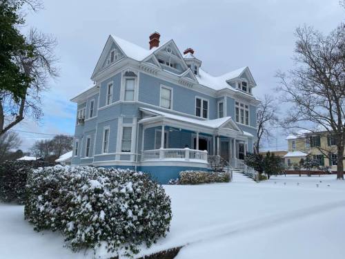A large blue Victorian house covered in snow, surrounded by white snow on the ground and bare trees.
