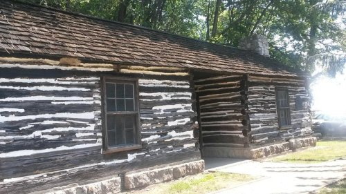 A rustic log cabin with a wooden exterior, featuring two windows and a stone foundation, surrounded by trees.