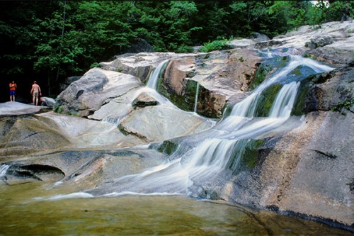 A serene waterfall cascades over smooth rocks, surrounded by lush greenery, with two people standing nearby.