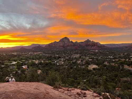 A stunning sunset over red rock formations and a valley, with two people sitting on a ledge in the foreground.