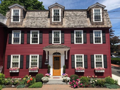 A charming red house with a shingled roof, black shutters, and flower boxes, surrounded by a well-kept garden.