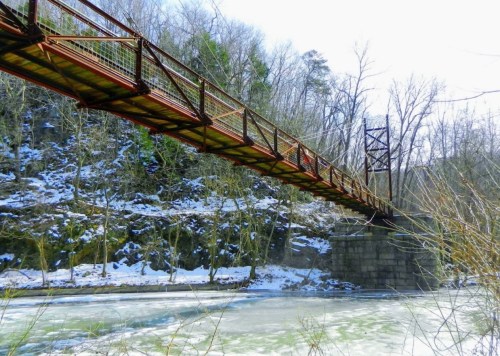 A suspension bridge spans a frozen river, surrounded by snow-covered trees and rocky cliffs.