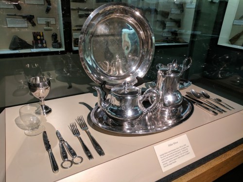 A display of silver tea set, tray, glassware, and cutlery in a museum exhibit.